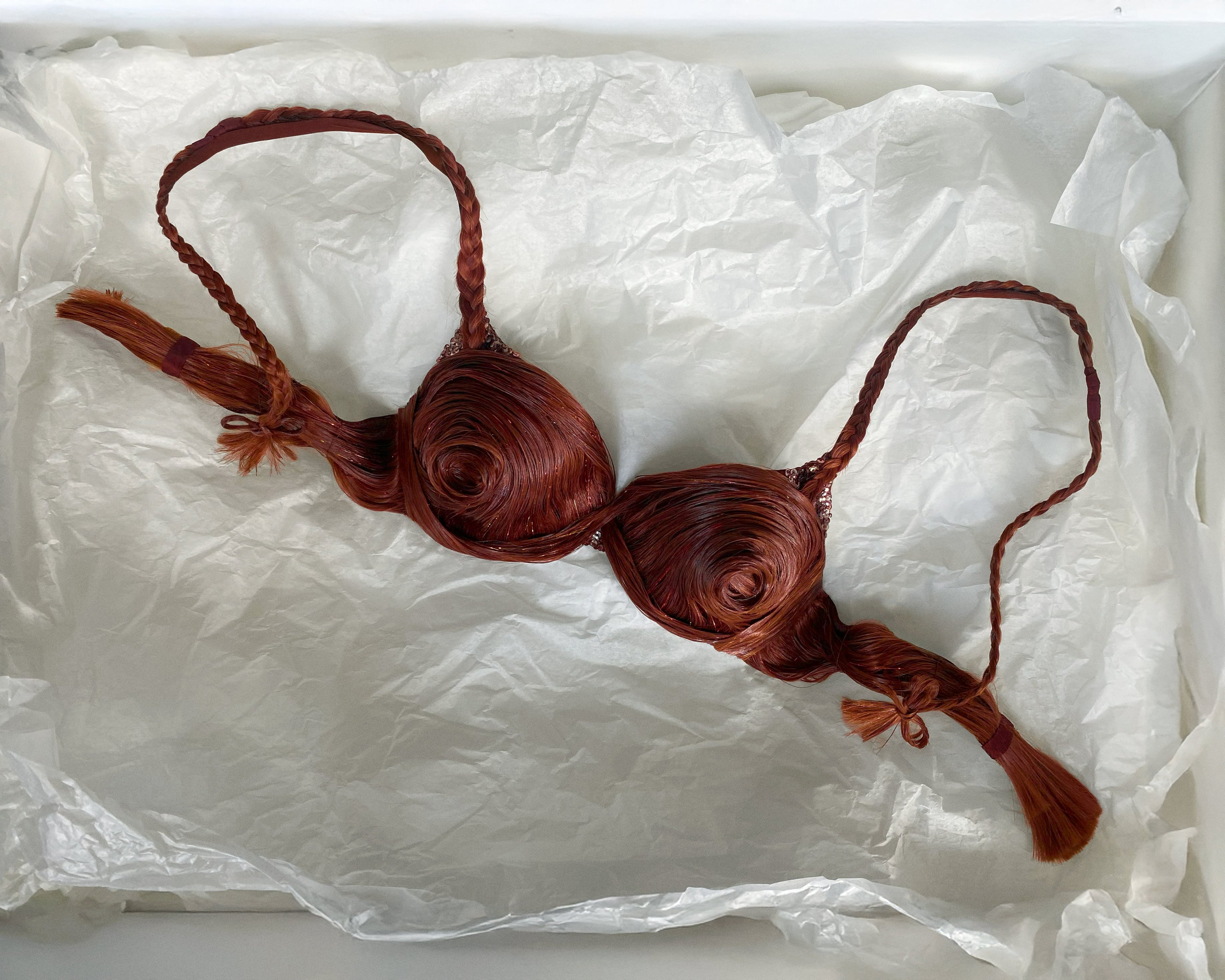 Custom demi-couture hair bra, designed and made by Australian fashion designer, Connor O'Grady. The bra is made from copper brown human hair, woven and embroidered with golden tinsel strands. In this image, the bra lays in a bed of white tissue paper.